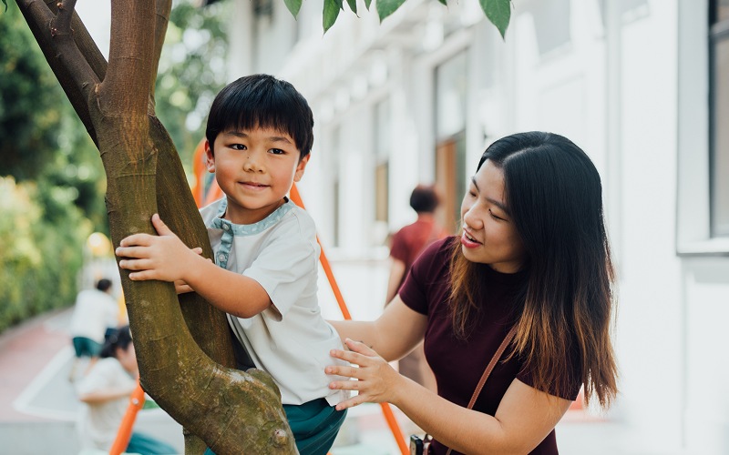 Teacher supervising a young boy climbing a tree outdoors.