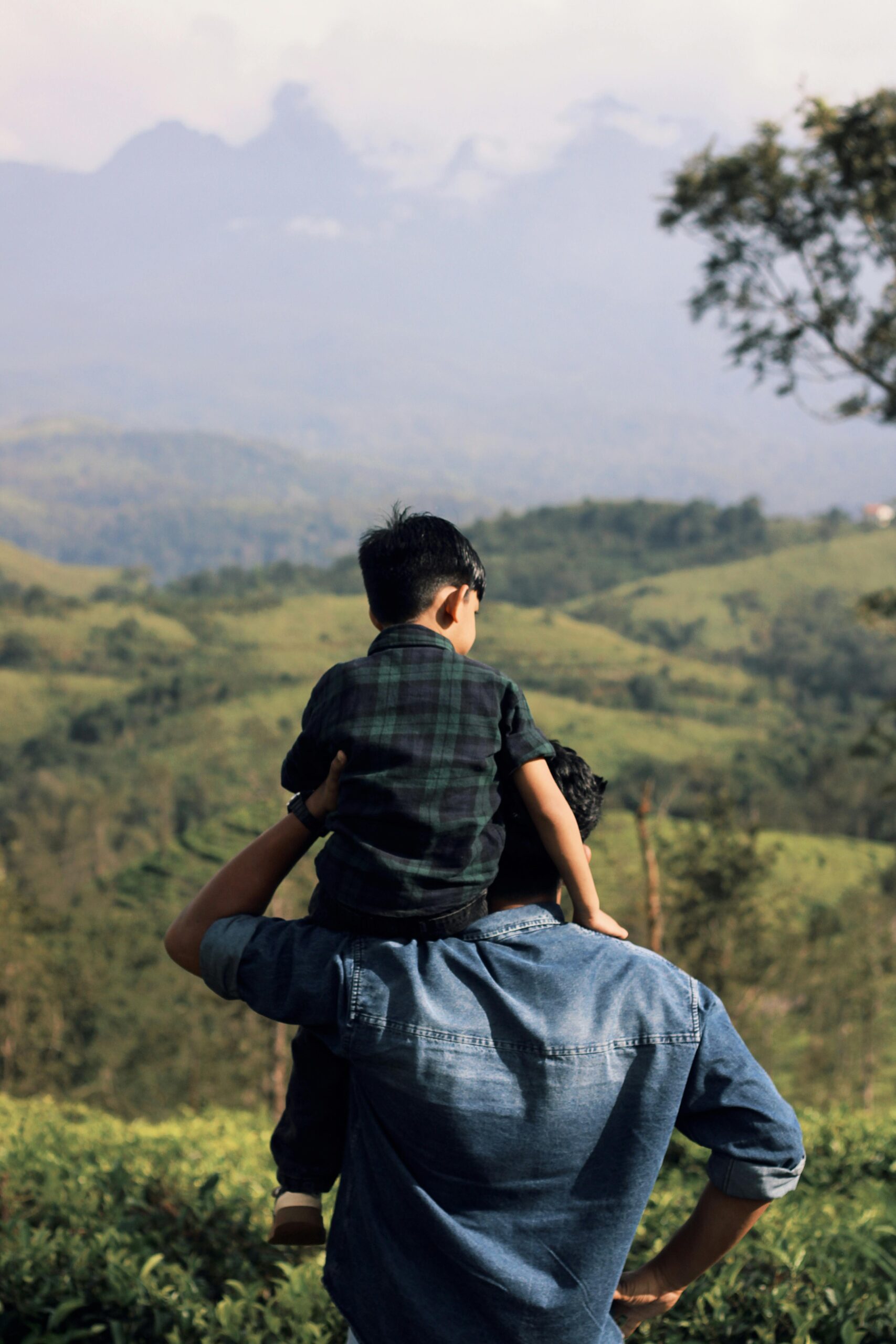 A father carrying his son on his shoulders, enjoying the lush greenery and mountainous landscape.