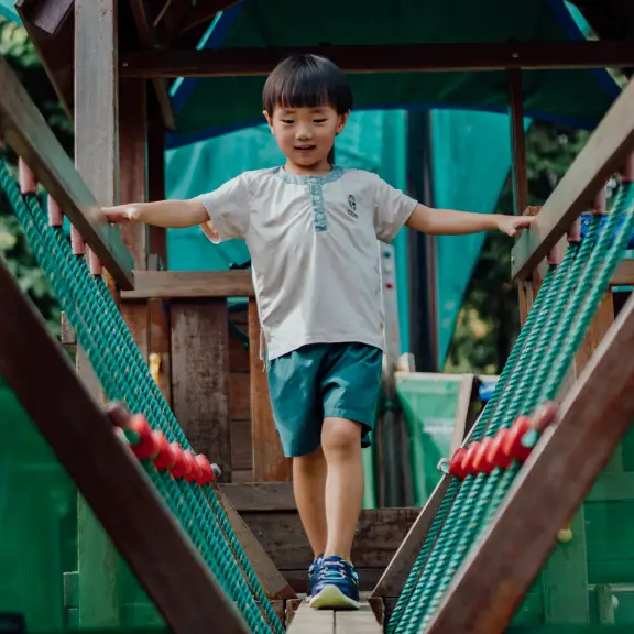 Young boy walking across a wooden rope bridge in play based preschool in Singapore